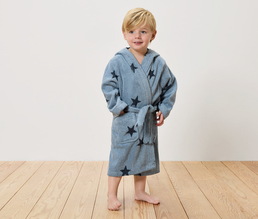 Malý chlapec s blond vlasy stojí bosý na dřevěné podlaze a má na sobě světle modrý župan s tmavými hvězdami.