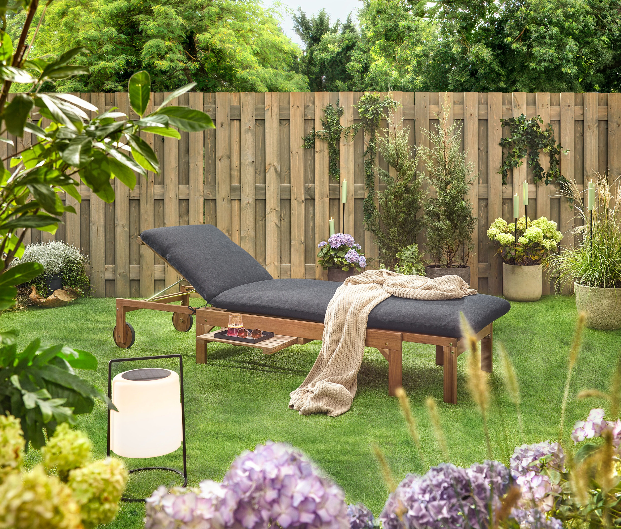 Zahradní scéna s dřevěným lehátkem s černým polstrováním, dekou, nápojem, slunečními brýlemi a lucernou na trávníku před dřevěným plotem.