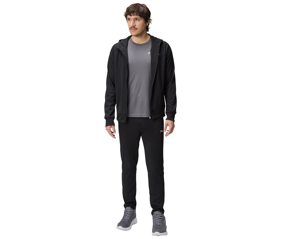 Muž v černé tréninkové bundě s kapucí, šedém tričku a černých teplácích a šedých sportovních botách.
