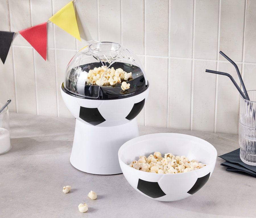 Stroj na popcorn ve tvaru fotbalového míče stojí vedle misky popcornu a sklenice s brčky.