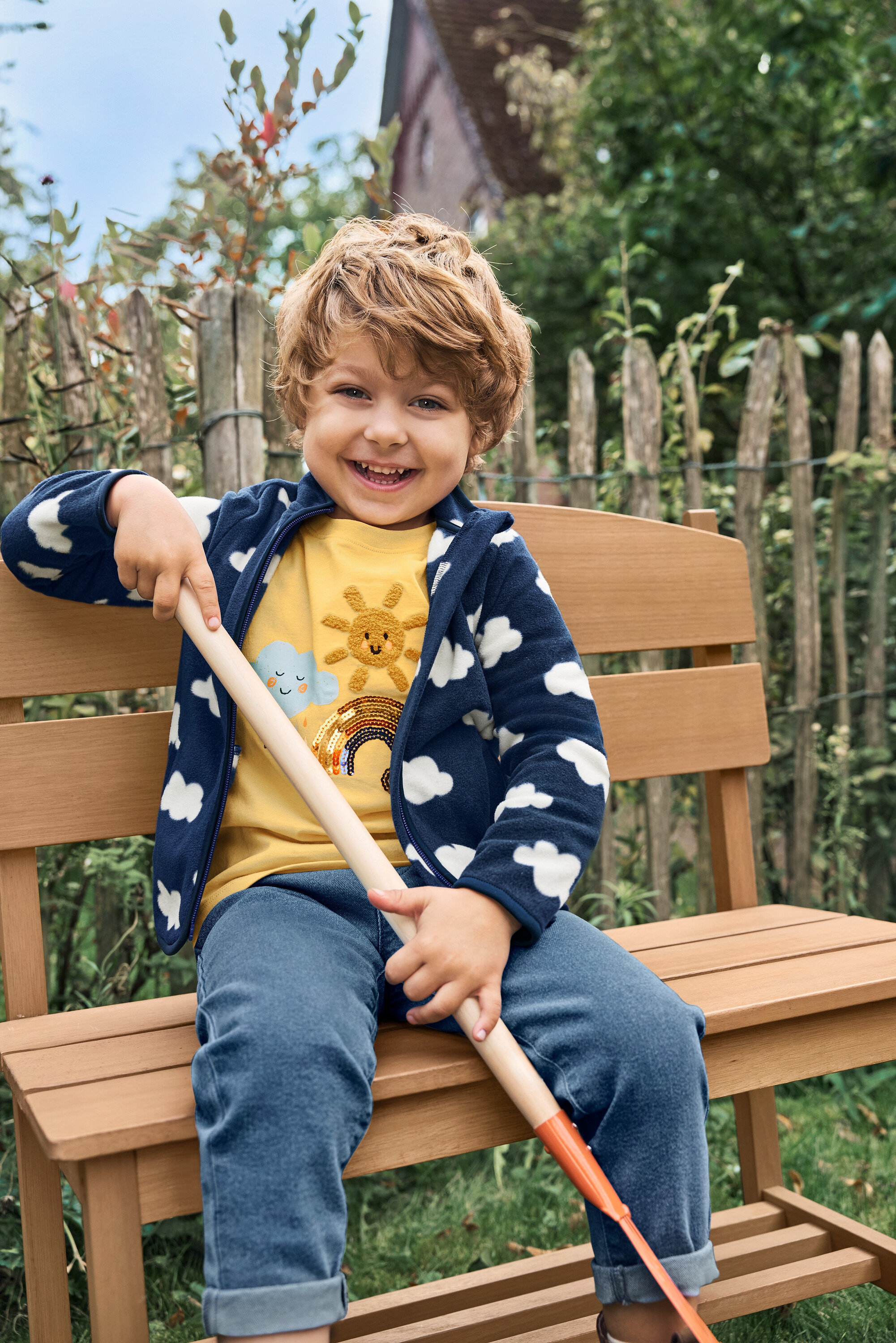 Usmívající se chlapec sedí na malé zahradní lavičce s přihrádkou a drží lopatu.
