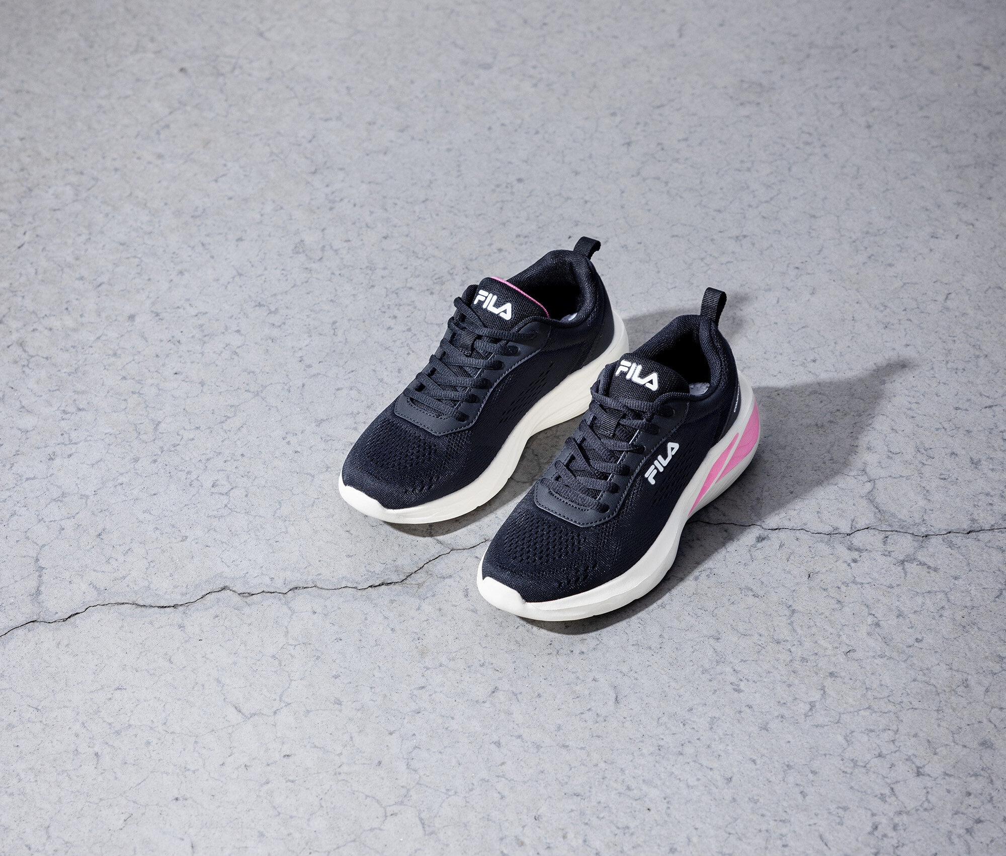 Dvě černé tenisky Fila leží na šedé betonové podlaze.
