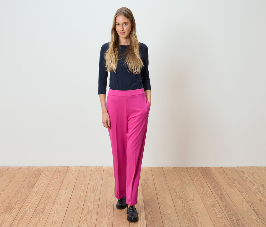 Žena stojí v tmavě modrém topu a růžových kalhotách před bílou stěnou.