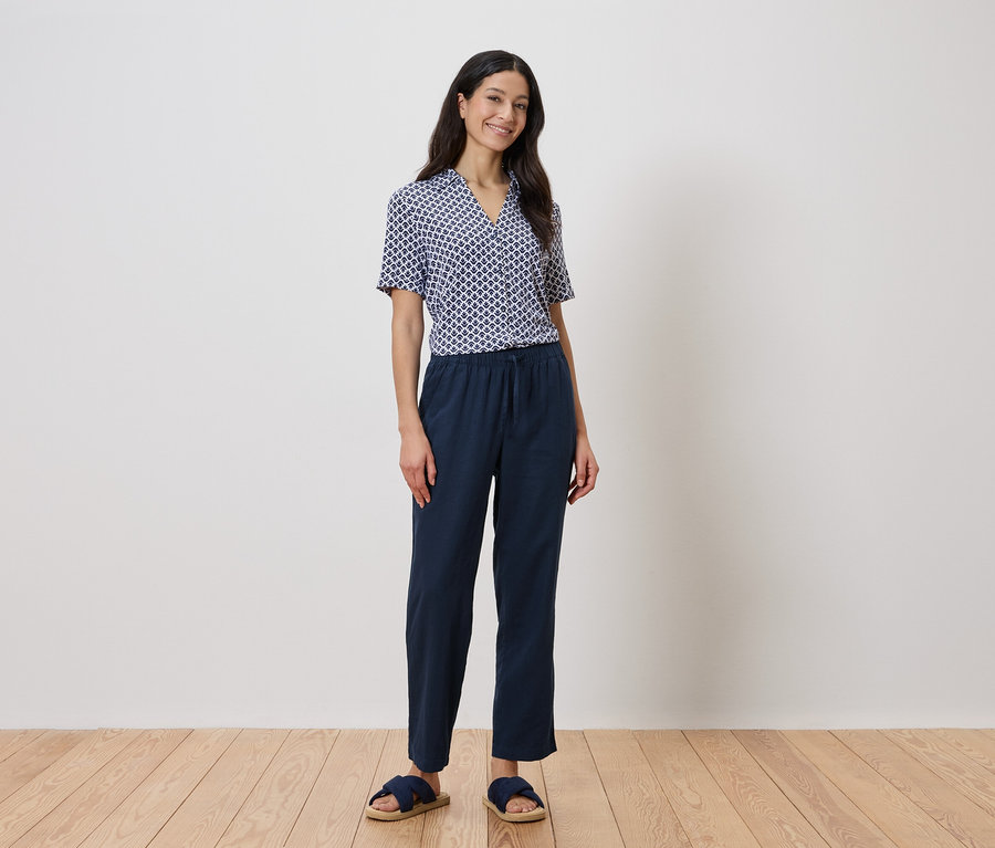 Žena stojí před stěnou a má na sobě modrou geometrickou košili a tmavě modré kalhoty.