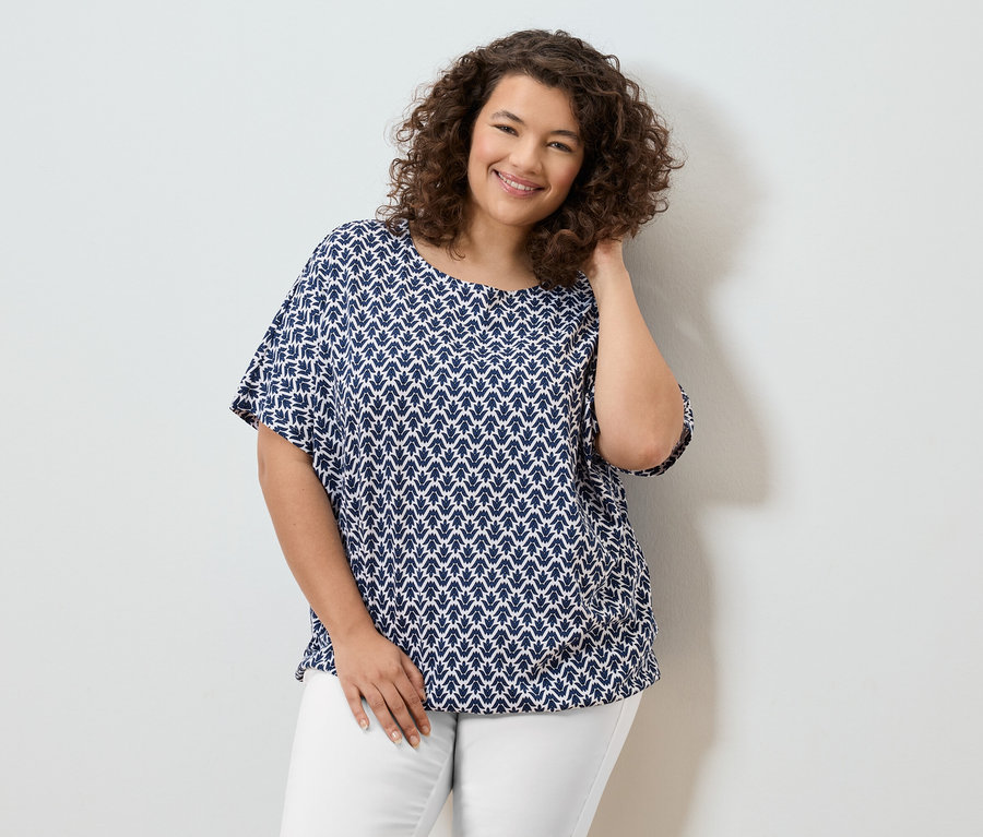 Usmívající se žena s kudrnatými vlasy v modrobílé halence a bílých kalhotách.