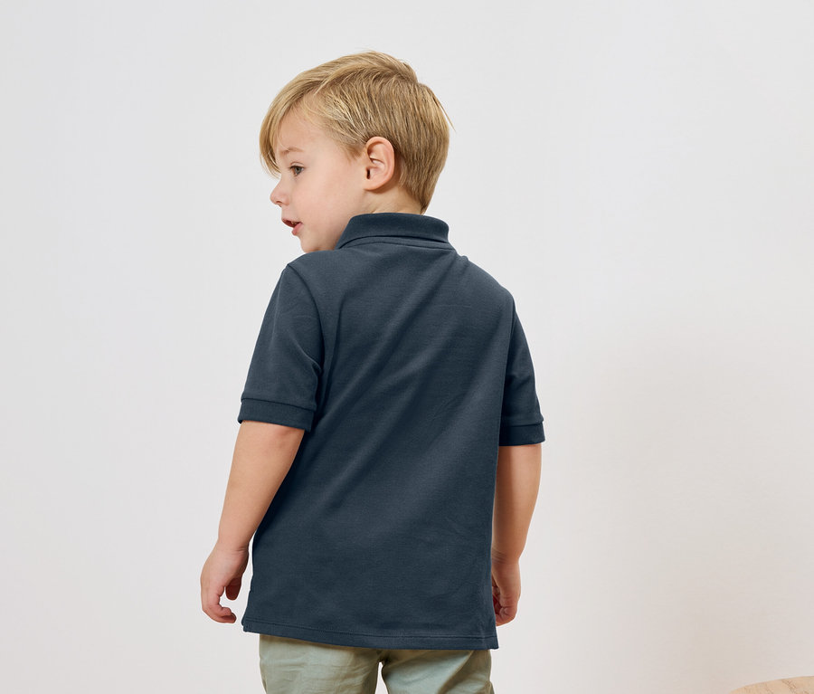 Chlapec s blond vlasy má na sobě tmavě modré polo tričko a světle zelené kalhoty, pohled zezadu.