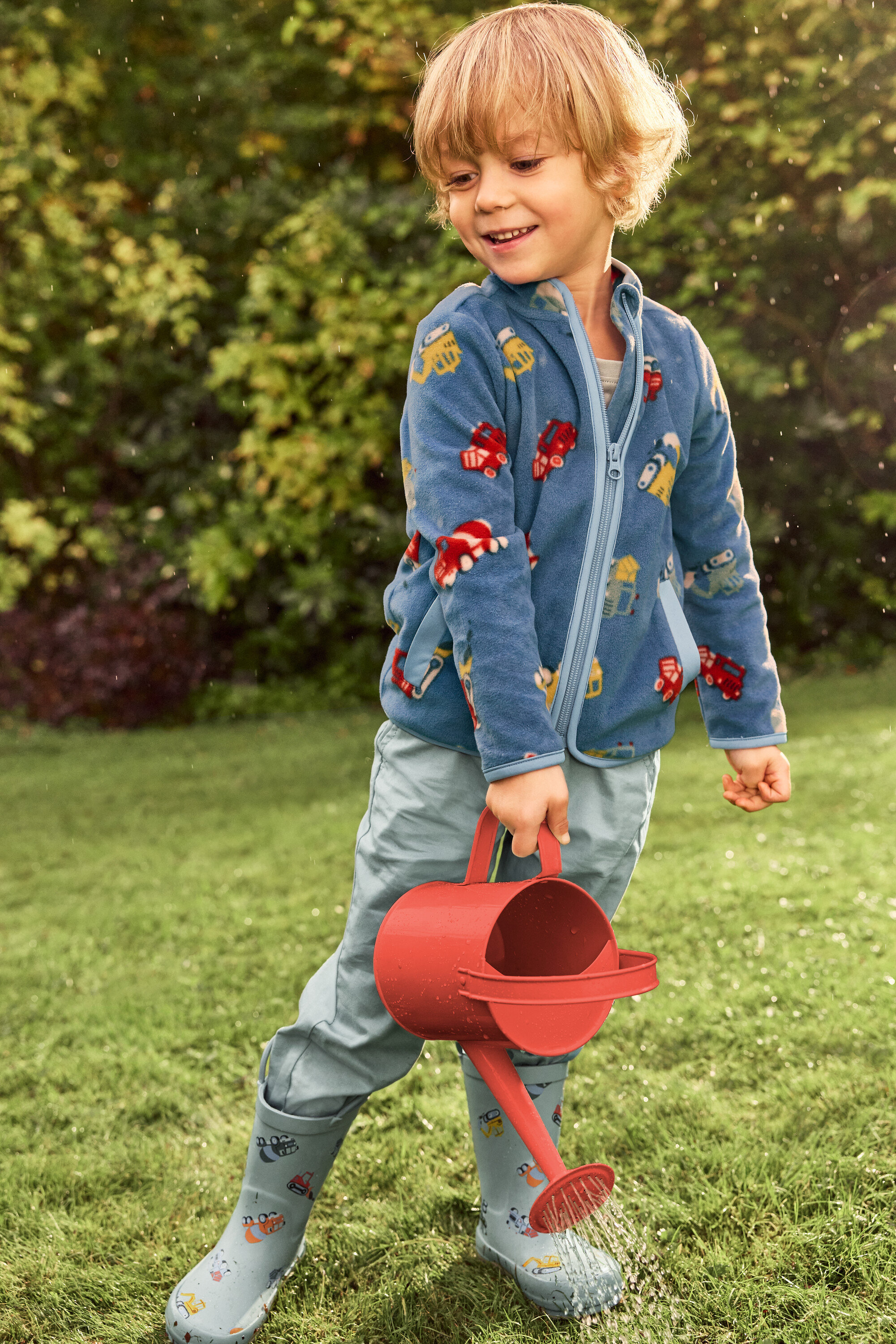 Usmívající se blonďatý chlapec zalévá červenou konev zelený trávník. Má na sobě modrou bundu s motivy vozidel a šedé gumáky s vozidly.