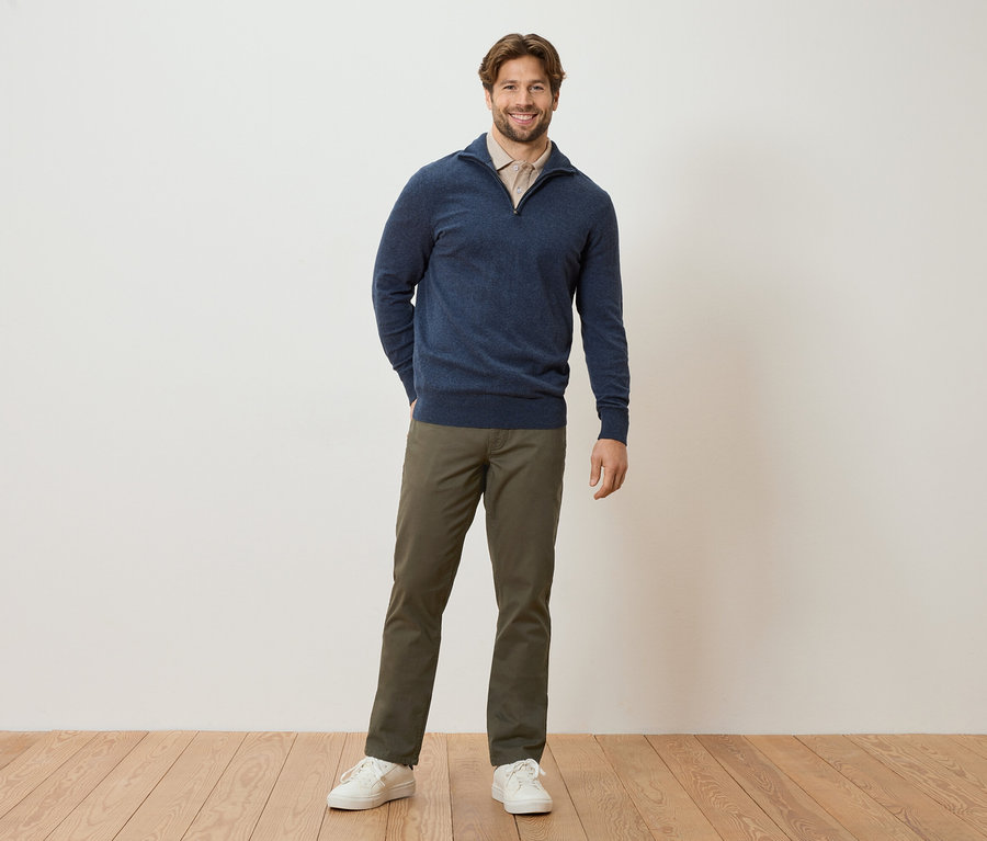 Muž stojí s rukama v kapsách před zdí. Má na sobě tmavě modrý svetr, světle hnědou blůzu, khaki kalhoty a bílé tenisky.