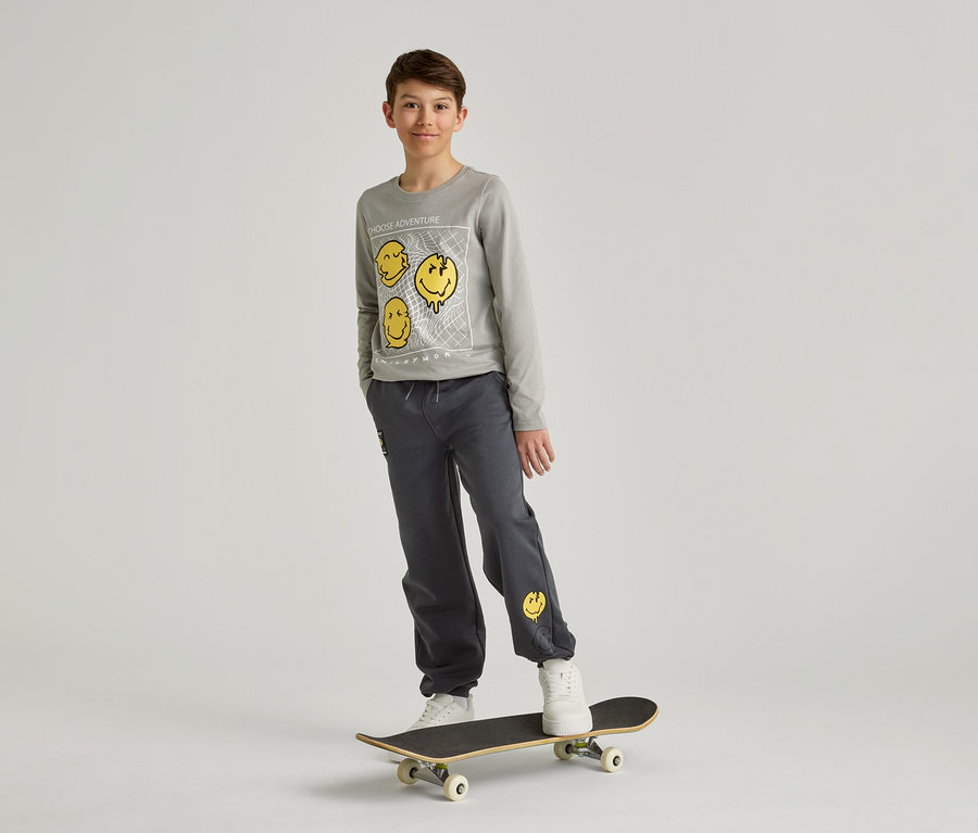 Chlapec v šedé mikině a kalhotách se žlutými smajlíky stojí na skateboardu před bílým pozadím.
