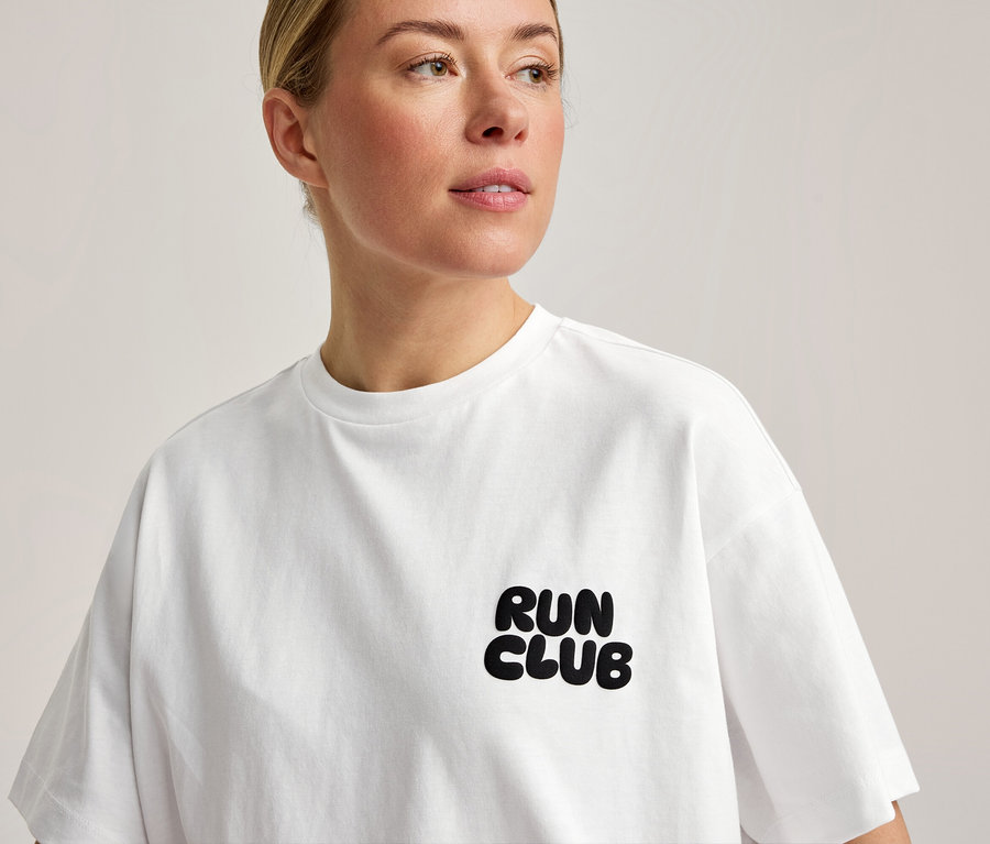 Žena má na sobě bílé sportovní tričko s nápisem „RUN CLUB“ na hrudi.
