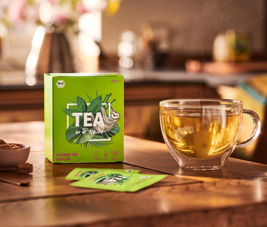Na dřevěném stole stojí balení BIO zeleného čaje TEA by Tchibo Sencha, miska s cukrem a šálek čaje.