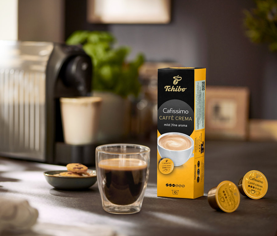 Na stole stojí kávovar, sklenice s kávou a balení Caffè Crema jemná – 120 kapslí.