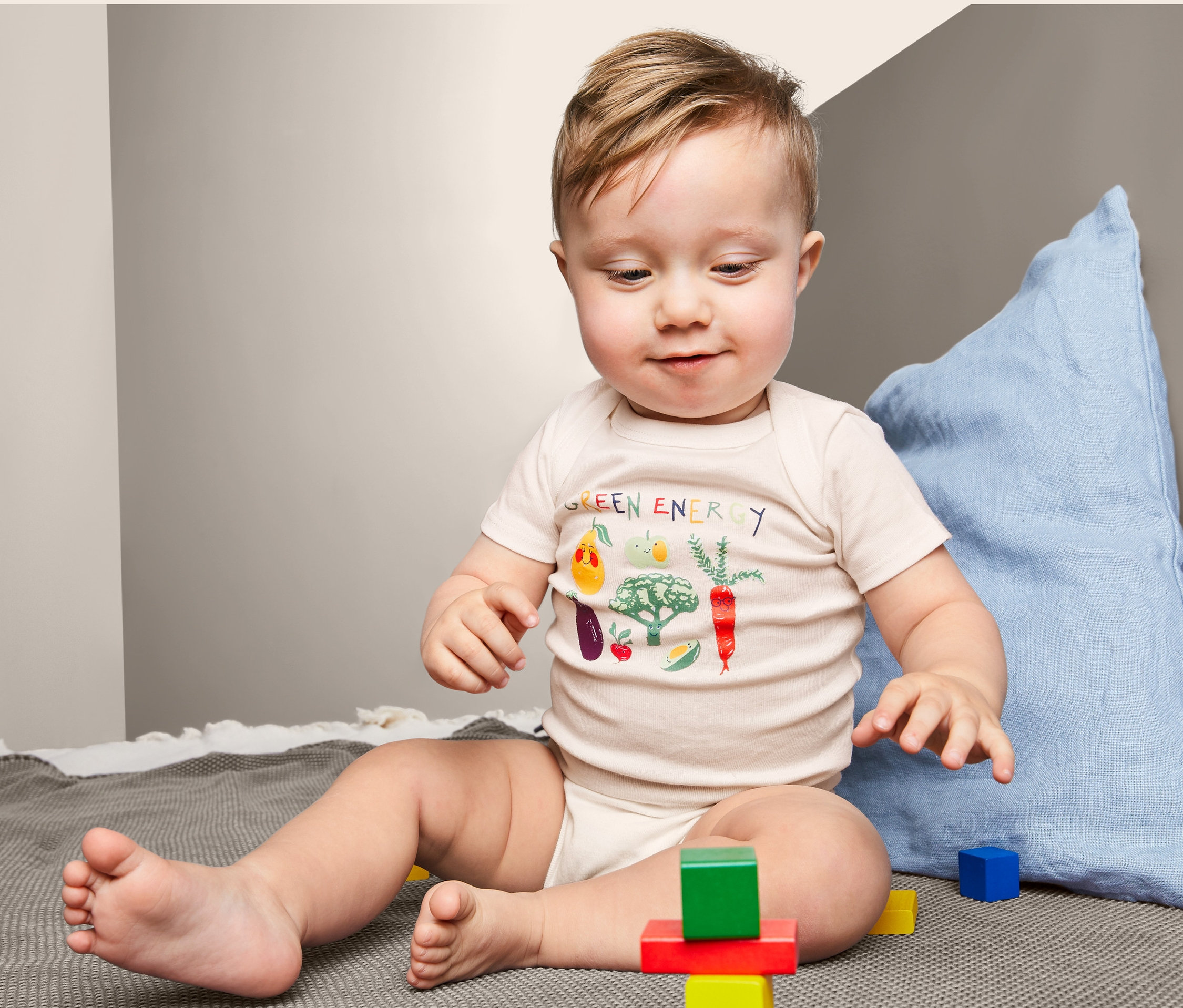 Dítě sedí na dece a hraje si s barevnými kostkami. Má na sobě body s krátkým rukávem s potiskem zeleniny.