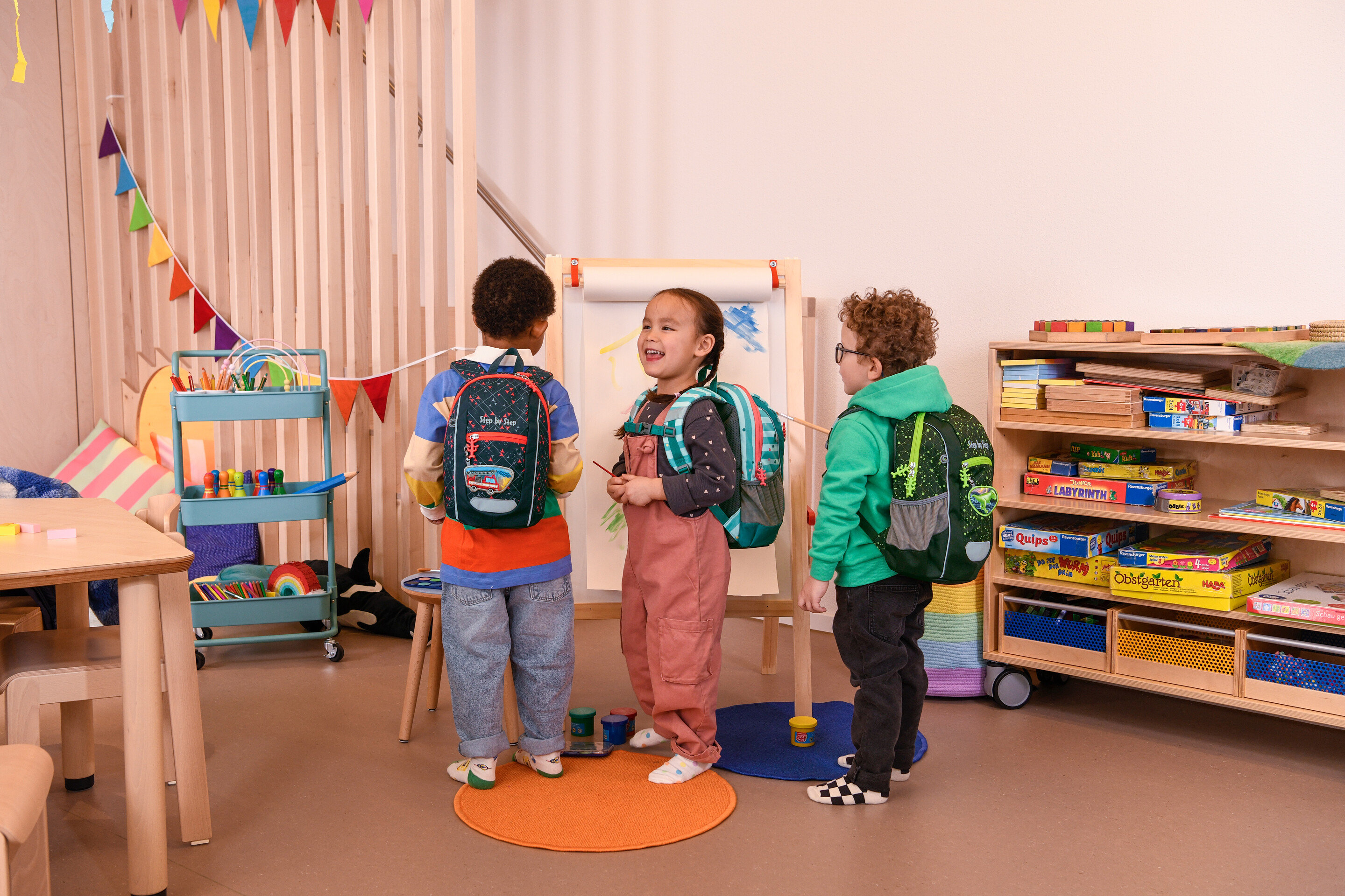 Tři děti stojí v místnosti s batohy a malují na plátno.