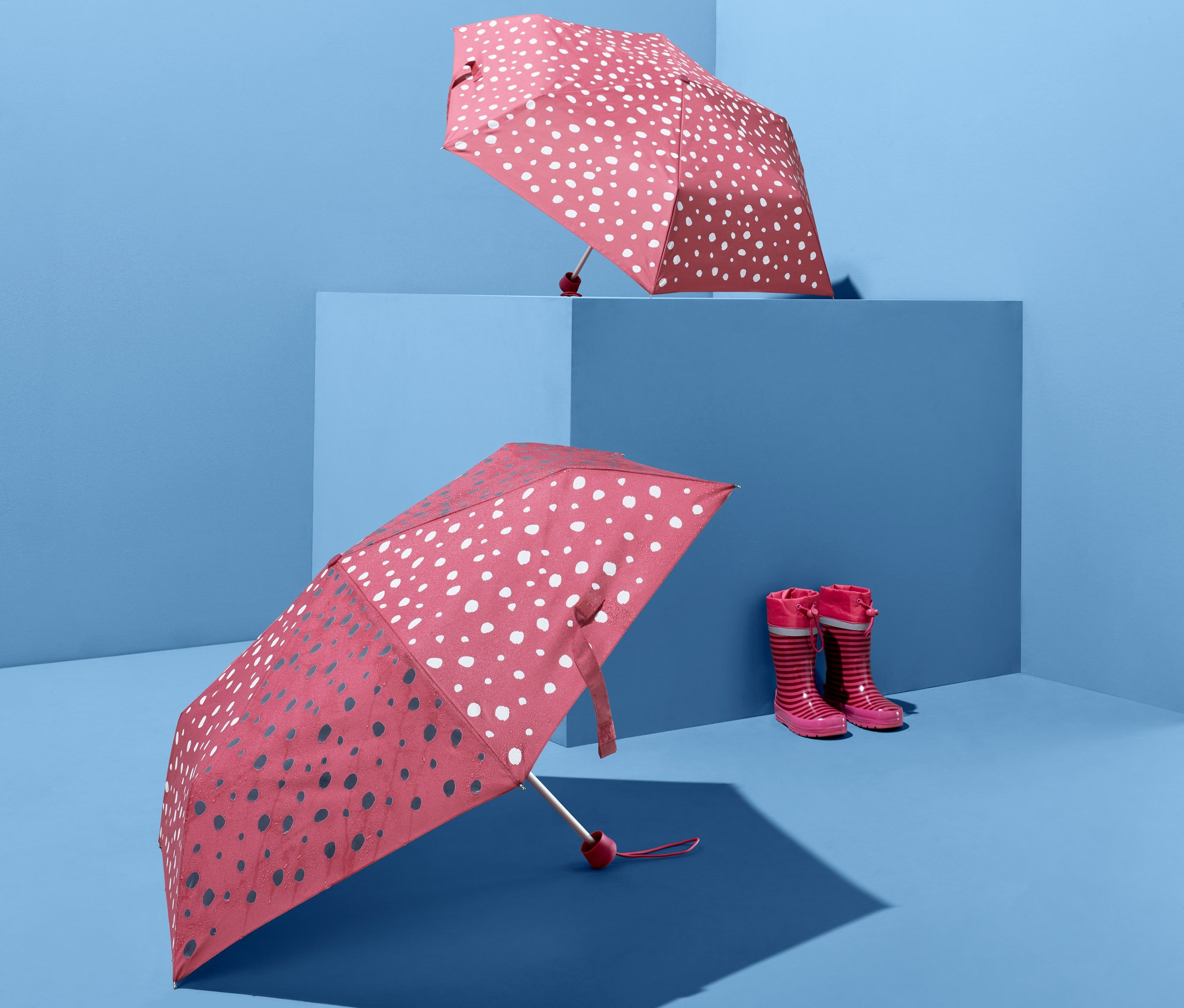 Dva dětské skládací deštníky s efektem změny barvy a pár dětských holínek na modrém pozadí.