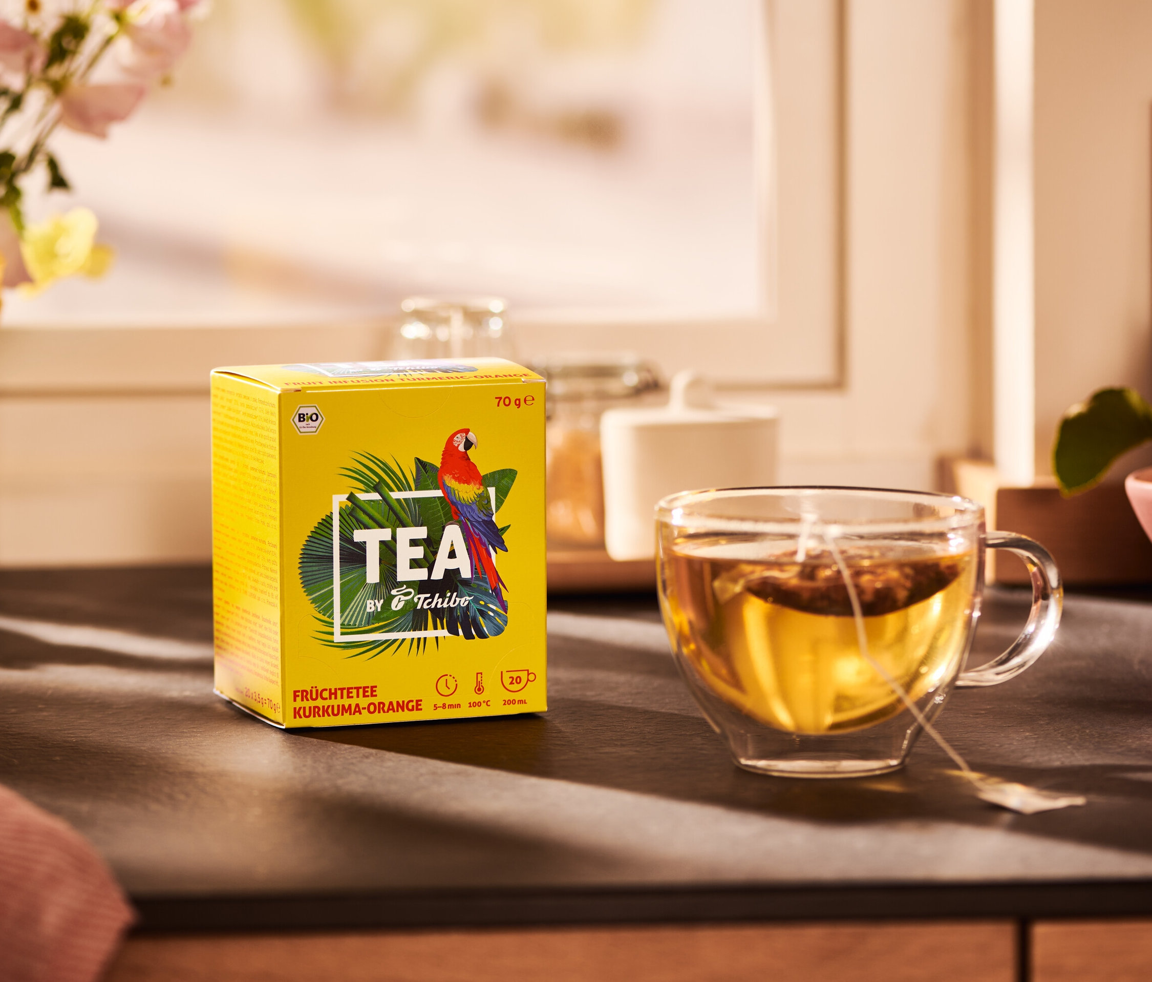 Žlutá krabička čaje TEA by Tchibo s příchutí kurkumy a pomeranče a šálek čaje.