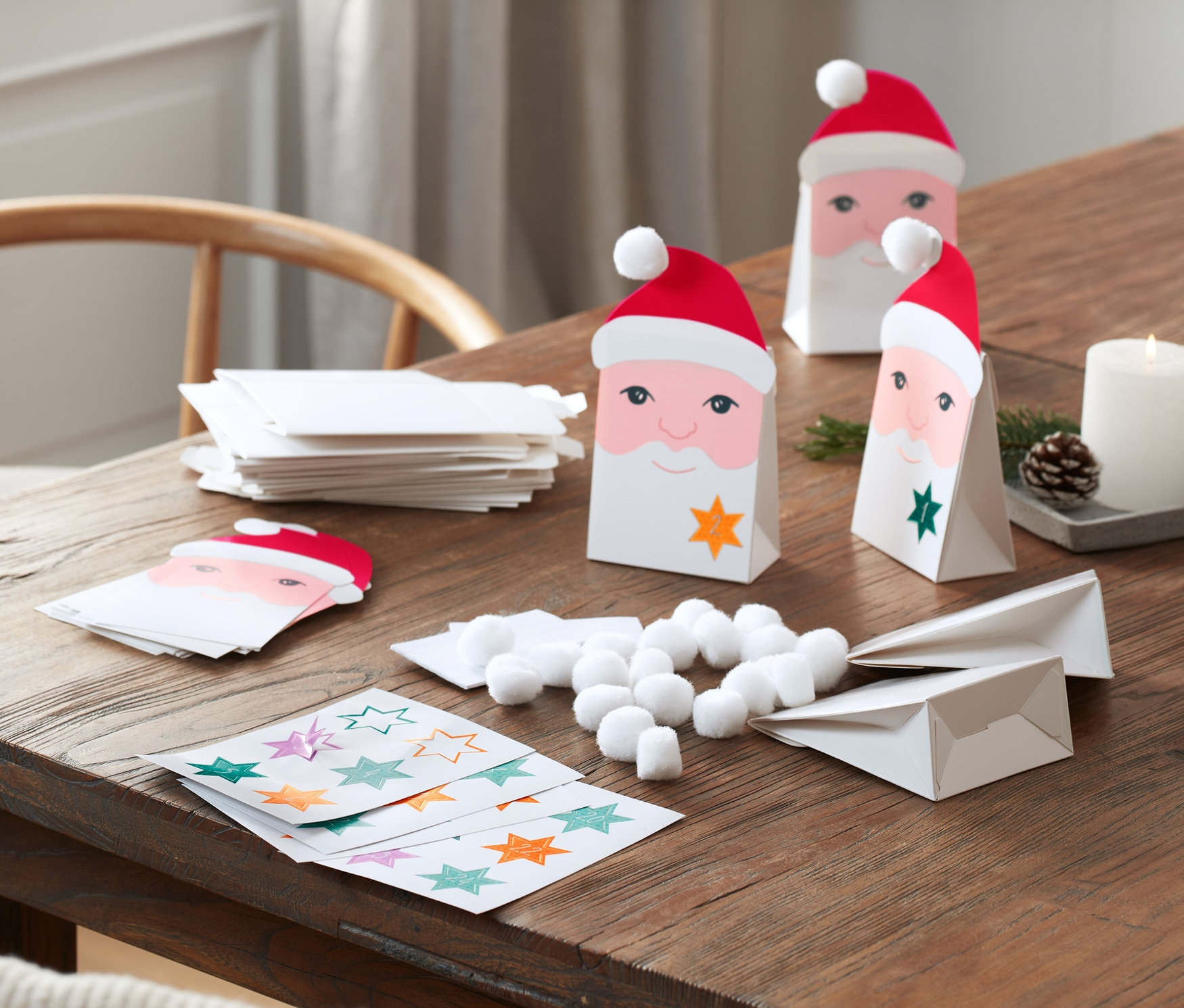 Na dřevěném stole leží sada na výrobu adventního kalendáře: papírové sáčky s obličejem Santy, samolepky s hvězdami, vatové kuličky a svíčky.