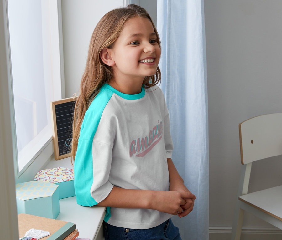 Dívka se usmívá a opírá o parapet v tričku z biobavlny s tyrkysovými rukávy.