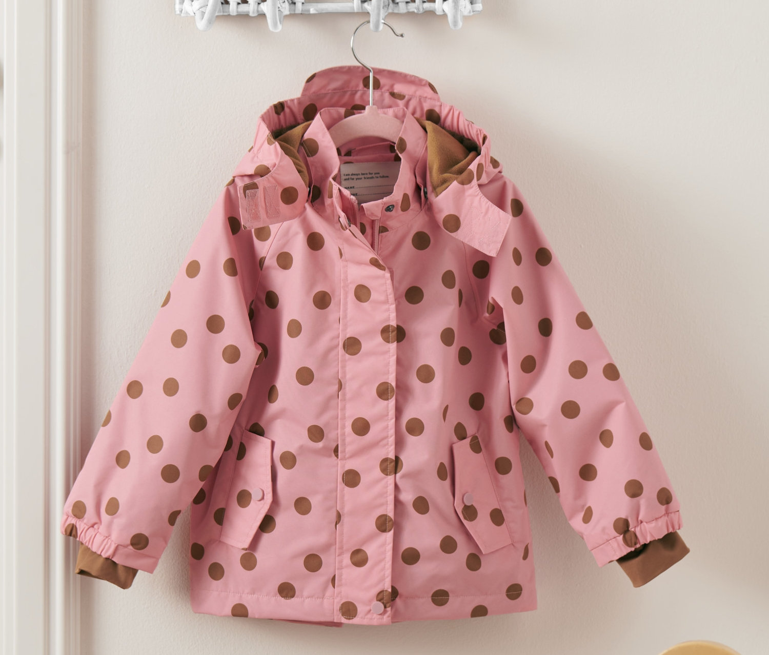Růžová termobunda do deště s hnědými puntíky visí na zdi vedle bílých dveří.