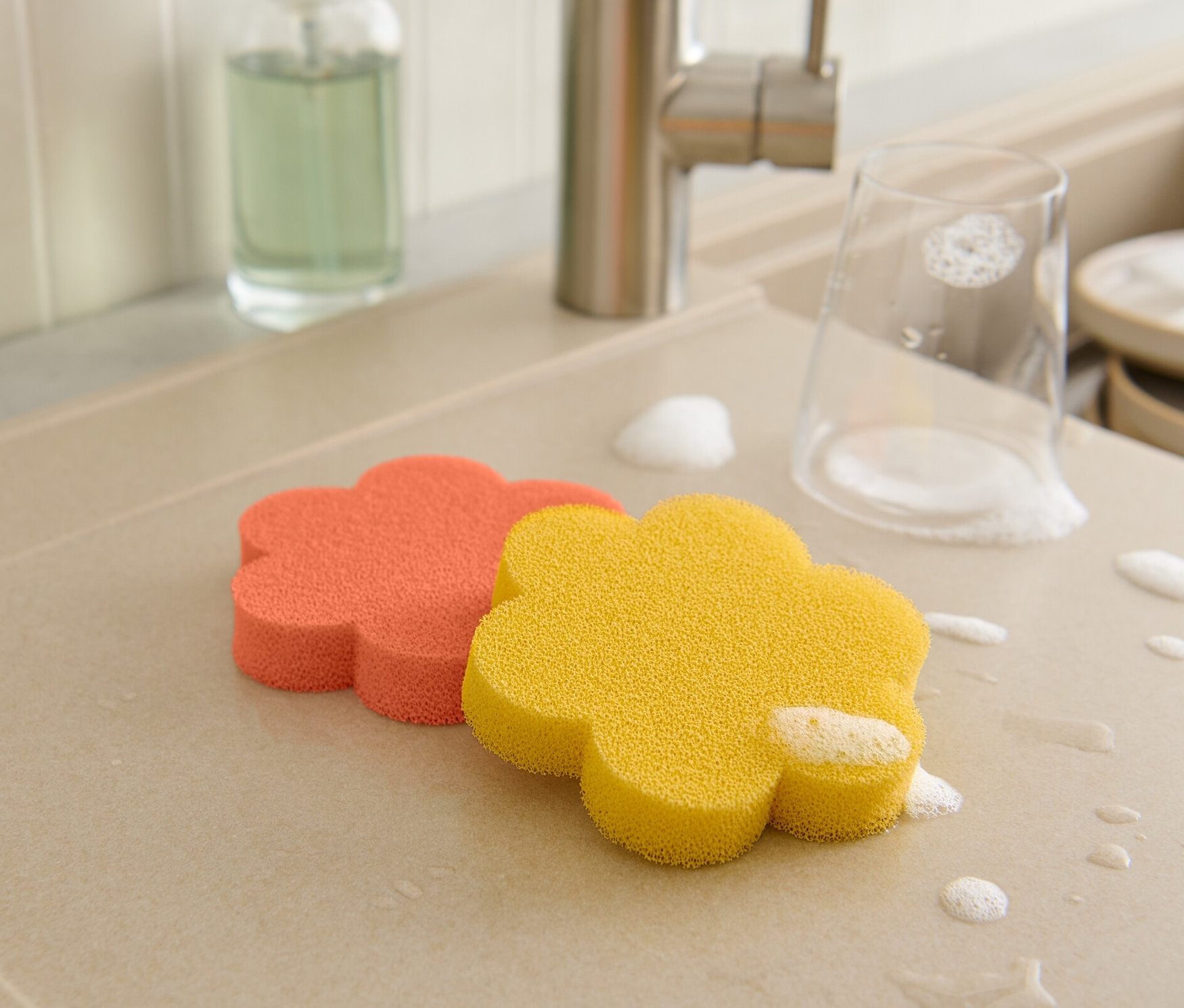 Na odkapávači leží oranžová a žlutá silikonová houbička ve tvaru květiny, sklenice a nádobí jsou pokryté pěnou.