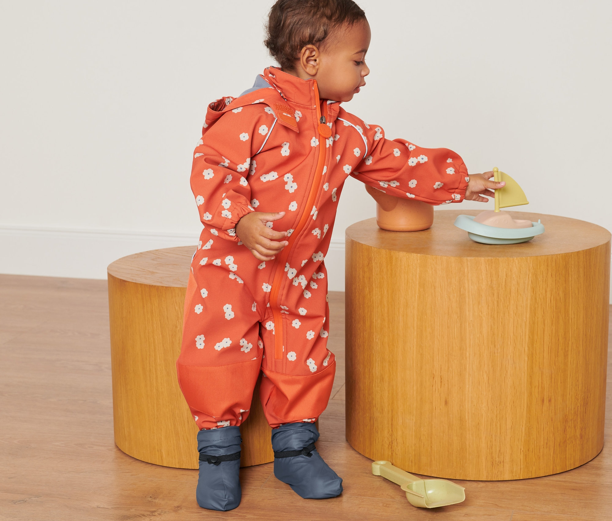 Dítě v oranžové kombinéze s květinovým vzorem si hraje s hračkou na dřevěném stolku. Má na sobě Ponožky do deště.