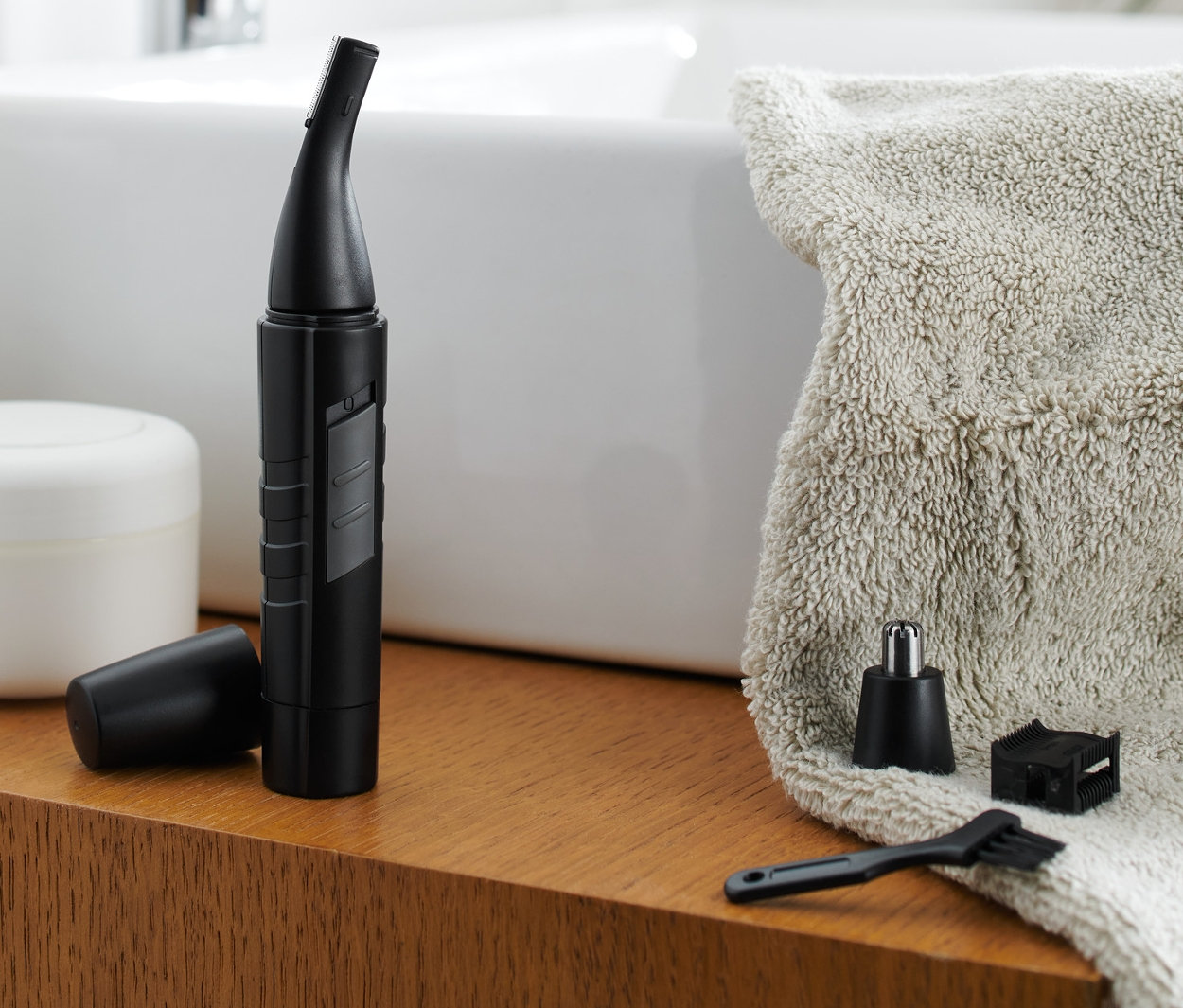 Na dřevěné polici v koupelně stojí zastřihovač chloupků v nose a uších spolu s příslušenstvím.