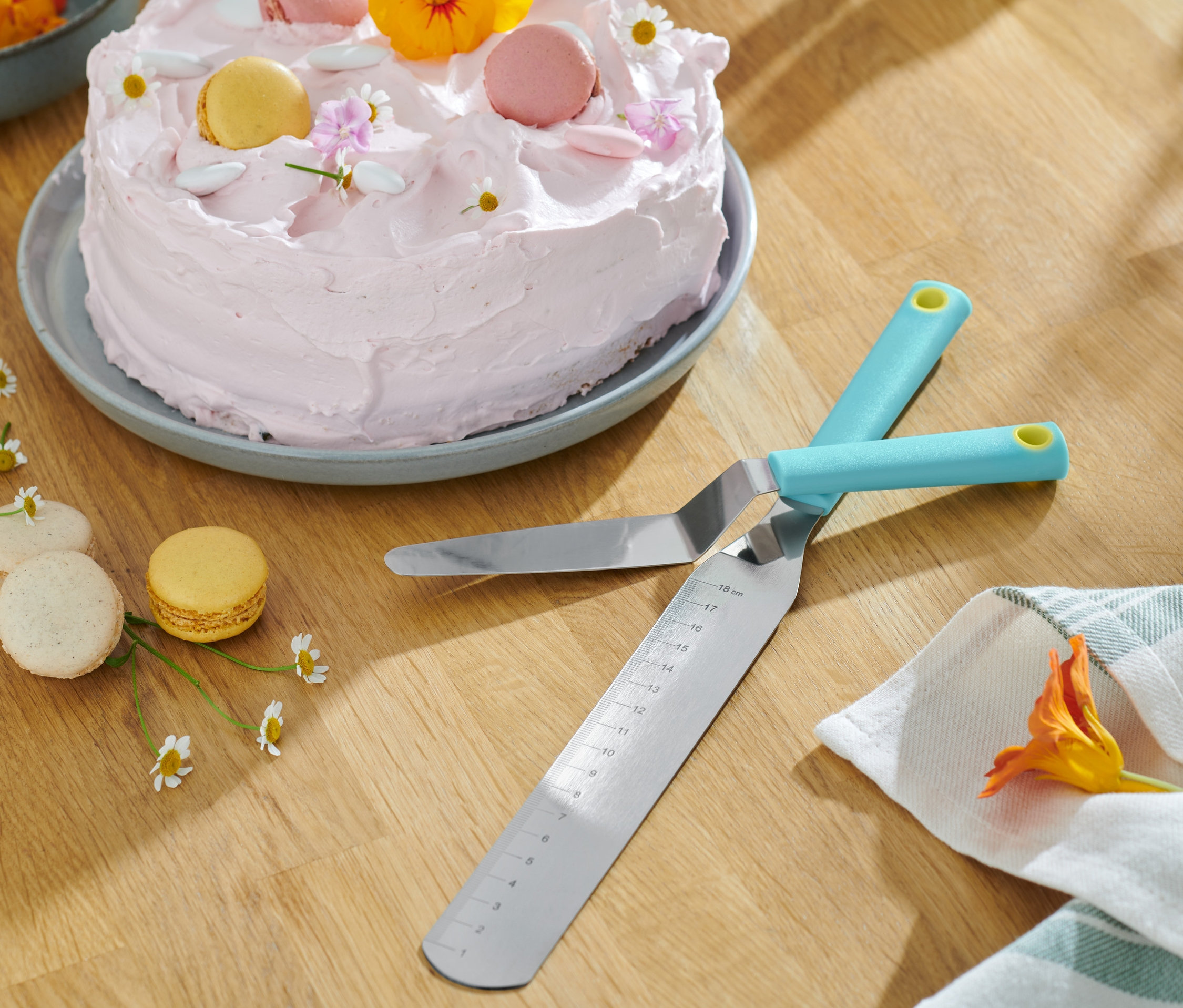 Dva roztírací nože leží zkřížené vedle sebe na dřevěném stole, v pozadí je dort s růžovým krémem a makronky.