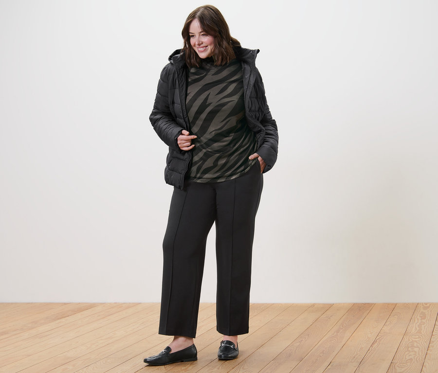 Žena pózuje v prošívané bundě s kapucí, síťovaném rolákovém triku s potiskem a žerzejových kalhotách s prošitím.