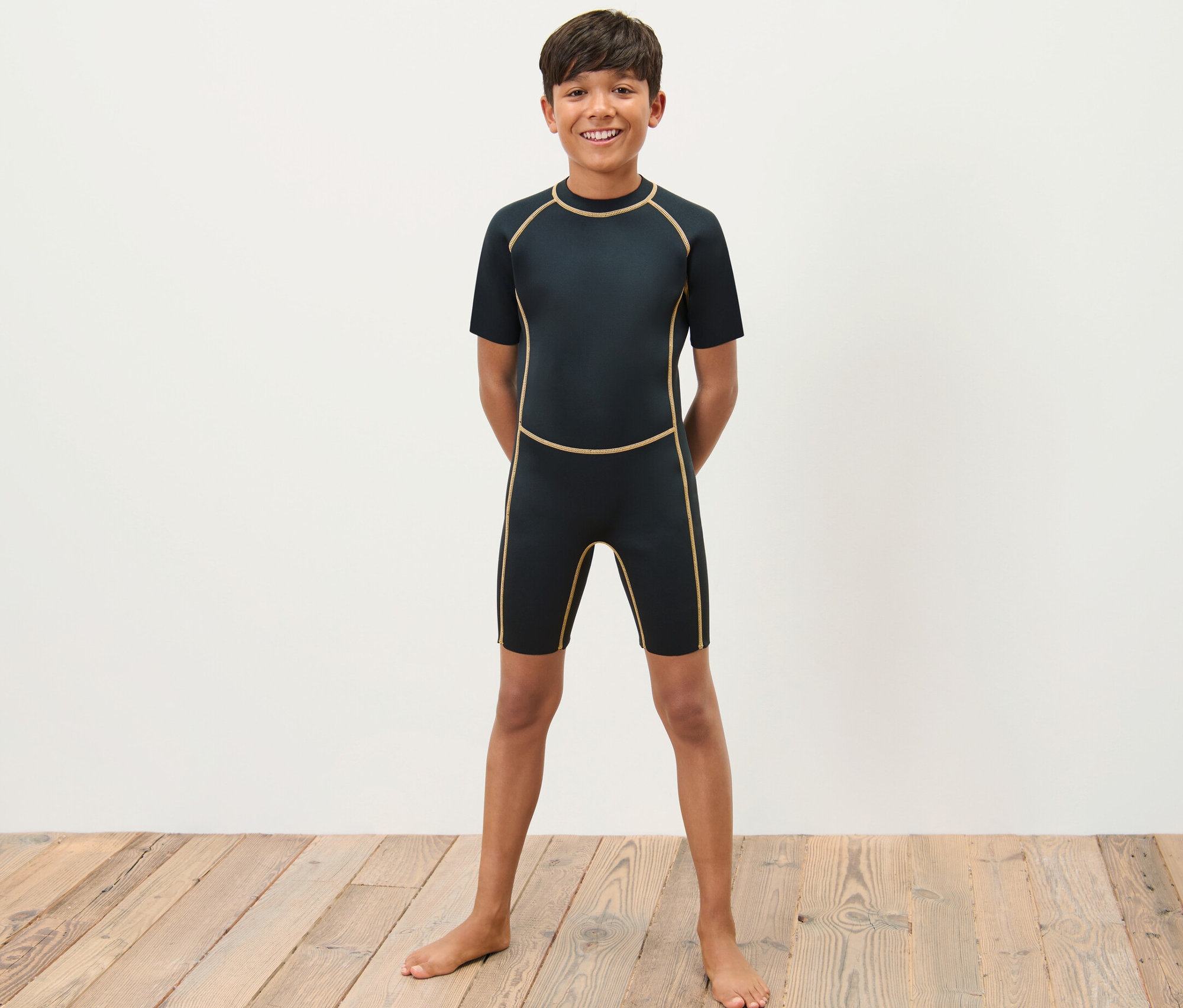 Chlapec v dětském neoprenovém obleku stojí bosý na dřevěné podlaze před bílou zdí.