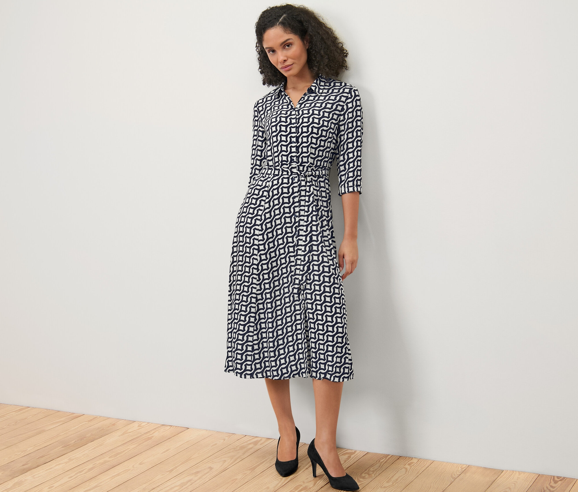 Žena stojící před bílou zdí má na sobě košilové šaty s potiskem a černé boty na podpatku.