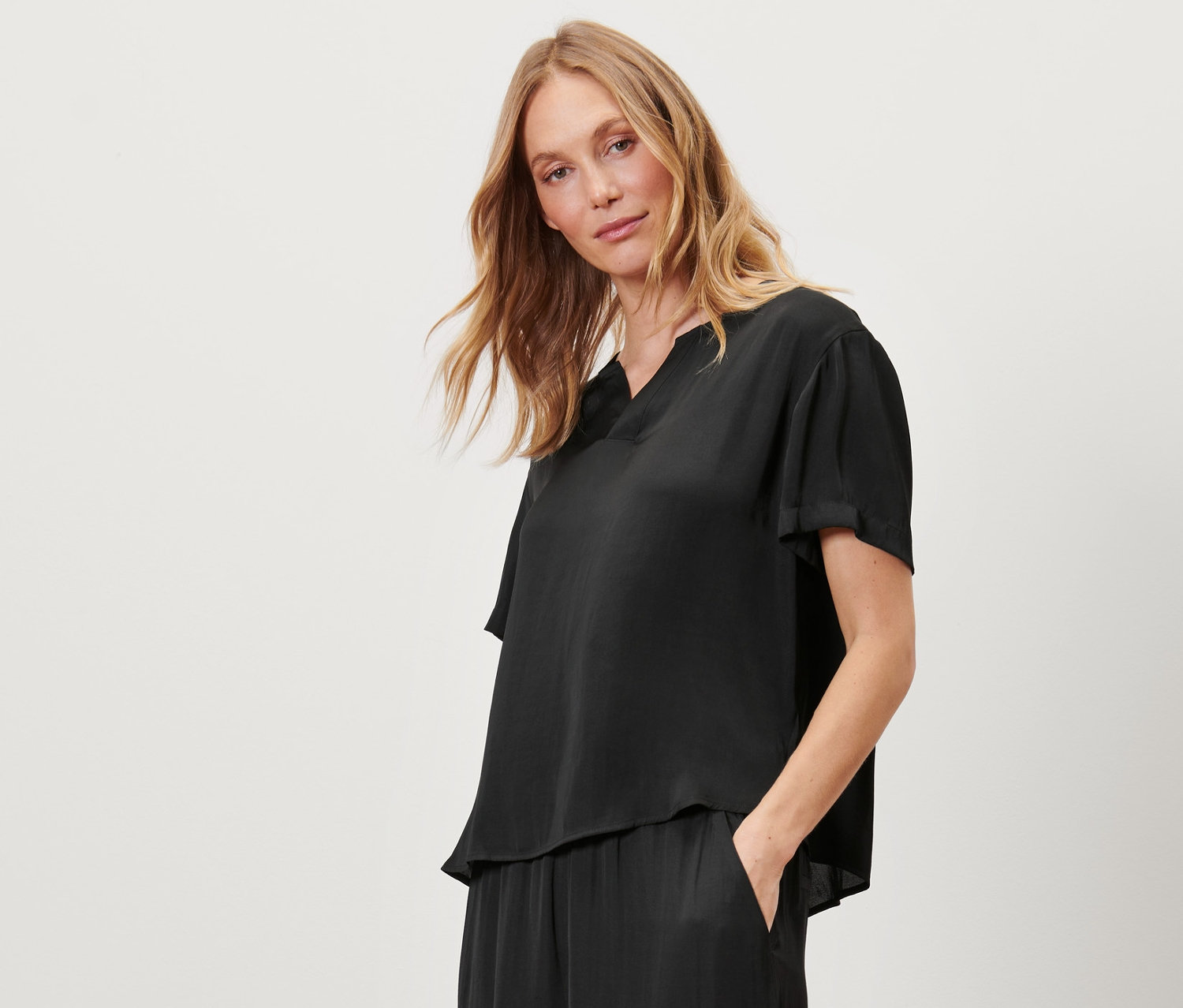 Žena stojí a má ruku v kapse saténové tunikové halenky, černé.