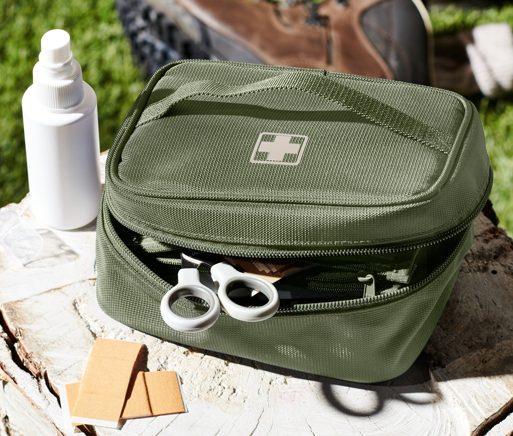 Zelená taštička na léky s bílým křížem leží otevřená na pařezu vedle bílé lahvičky s pumpičkou a náplastí.