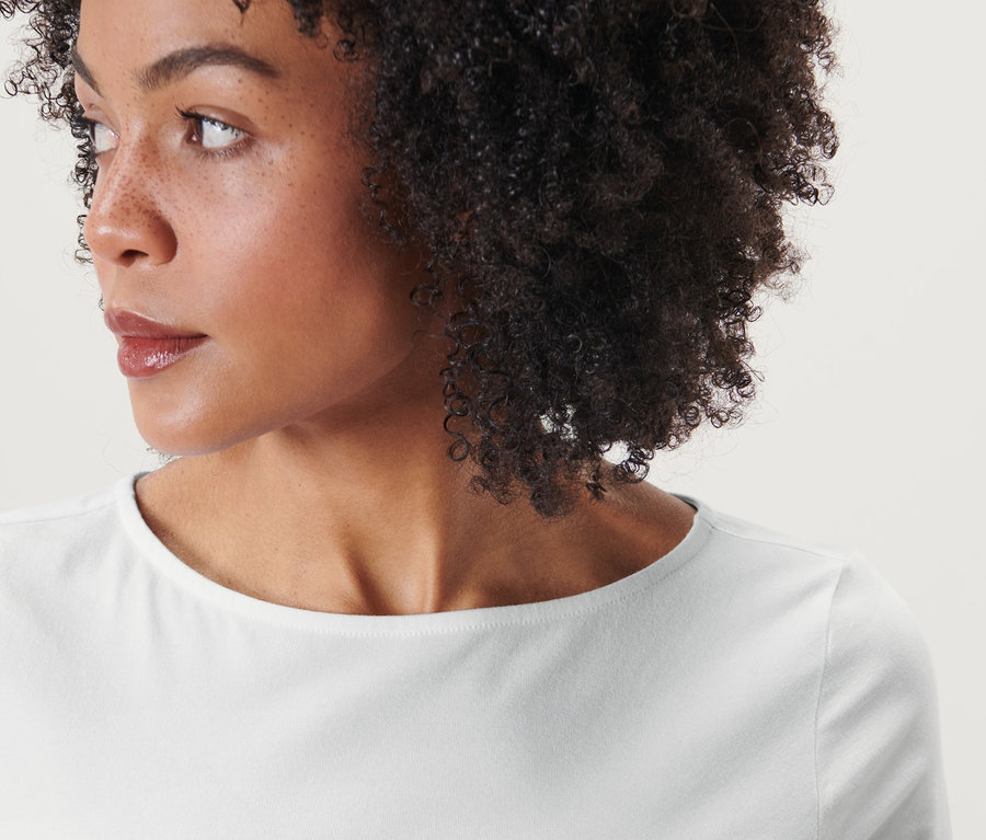 Portrét ženy s hnědými kudrnatými vlasy, která má na sobě bílé triko s tříčtvrtečním rukávem.