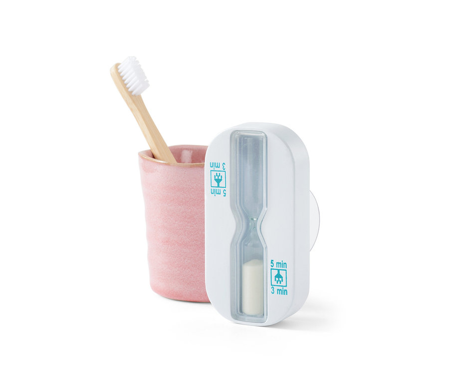 Bambusový zubní kartáček v růžovém kelímku vedle bílých přesýpacích hodin do sprchy.