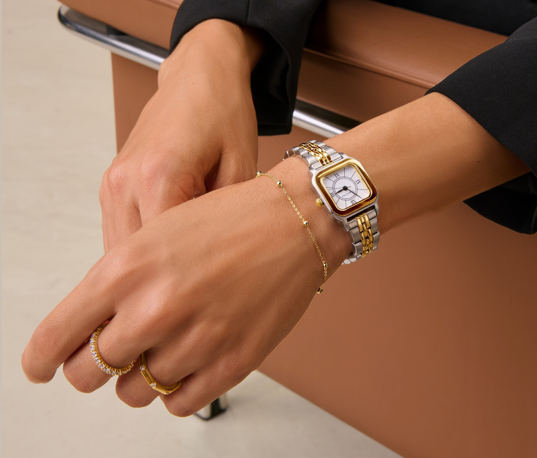 Žena sedí na hnědé kožené židli a má na sobě dvoubarevné dámské hodinky z ušlechtilé oceli, zlatý kuličkový náramek a dva zlaté prsteny.