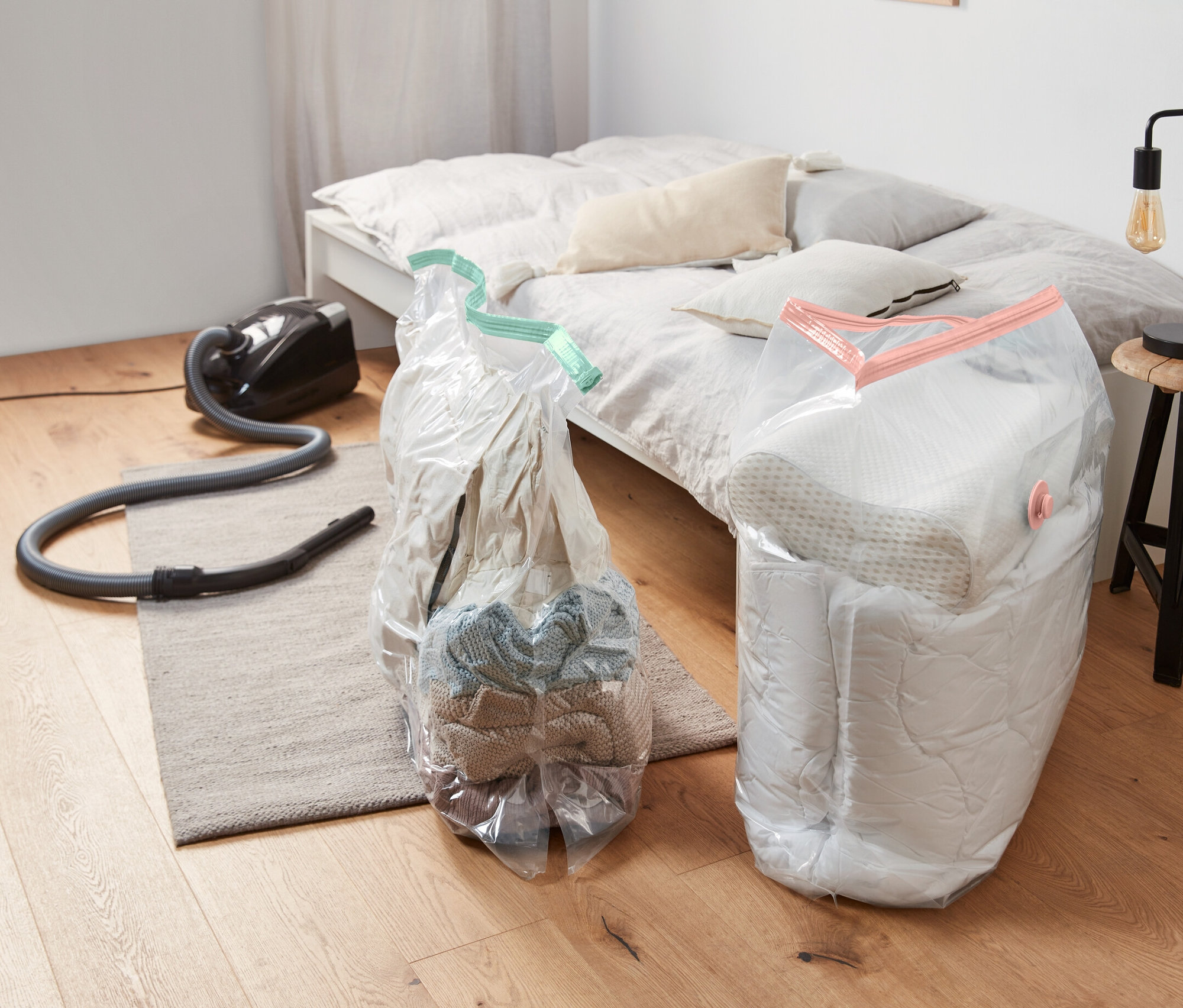 Dva vakuové úložné pytle, 2 ks, hranaté, jsou umístěny na podlaze vedle vysavače a postele.