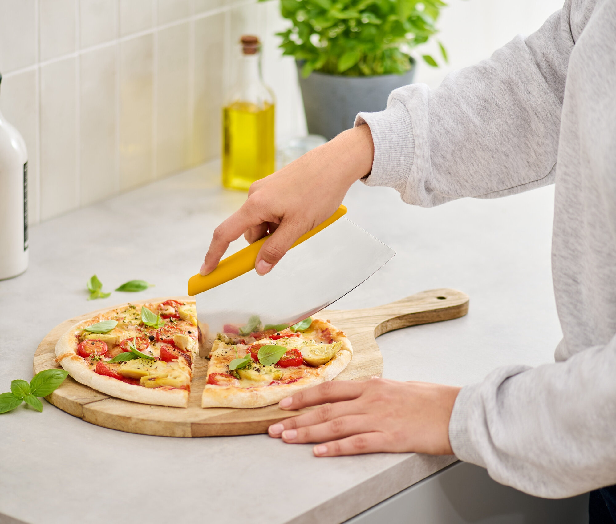 Žena krájí pizzu s rajčaty a bazalkou na dřevěném prkénku žlutým kráječem na lince.