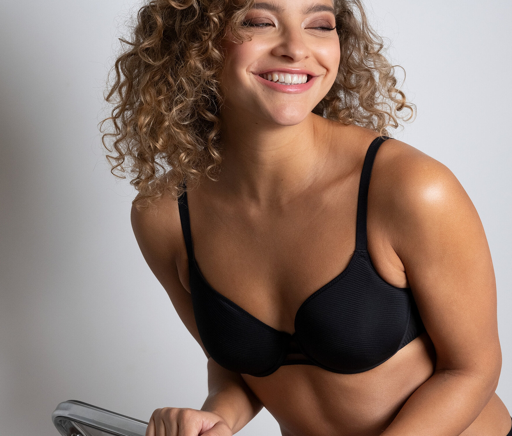 Usmívající se žena sedí na průhledné židli a má na sobě černou podprsenku Passionata »Nais«.