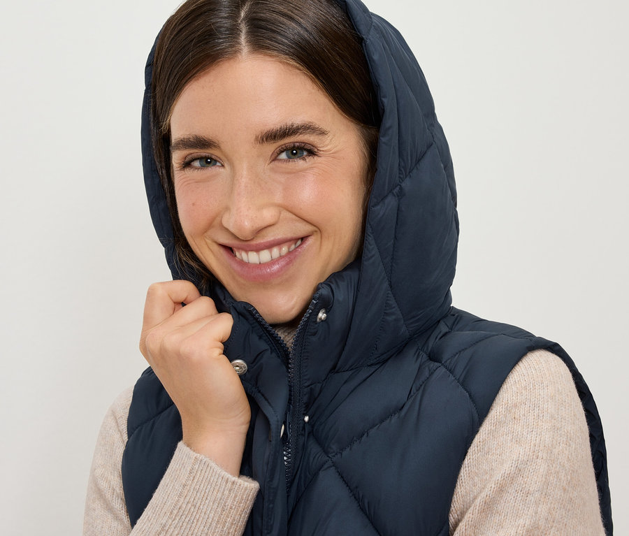 Usmívající se žena má na sobě tmavě modrou prošívanou vestu s kapucí.
