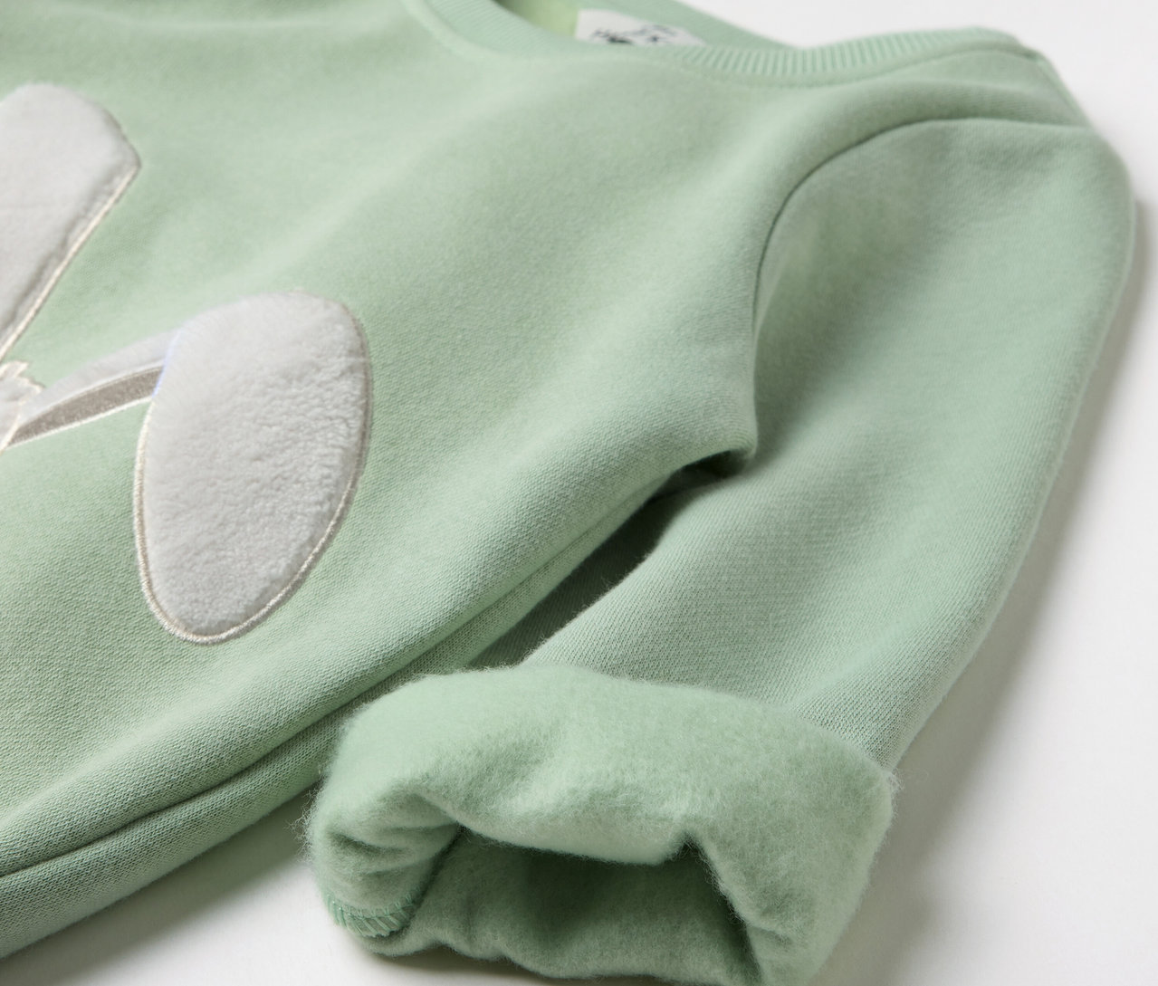 Světlý zelený svetr s nášivkou králičích uší na hrudi a ohrnutými rukávy.