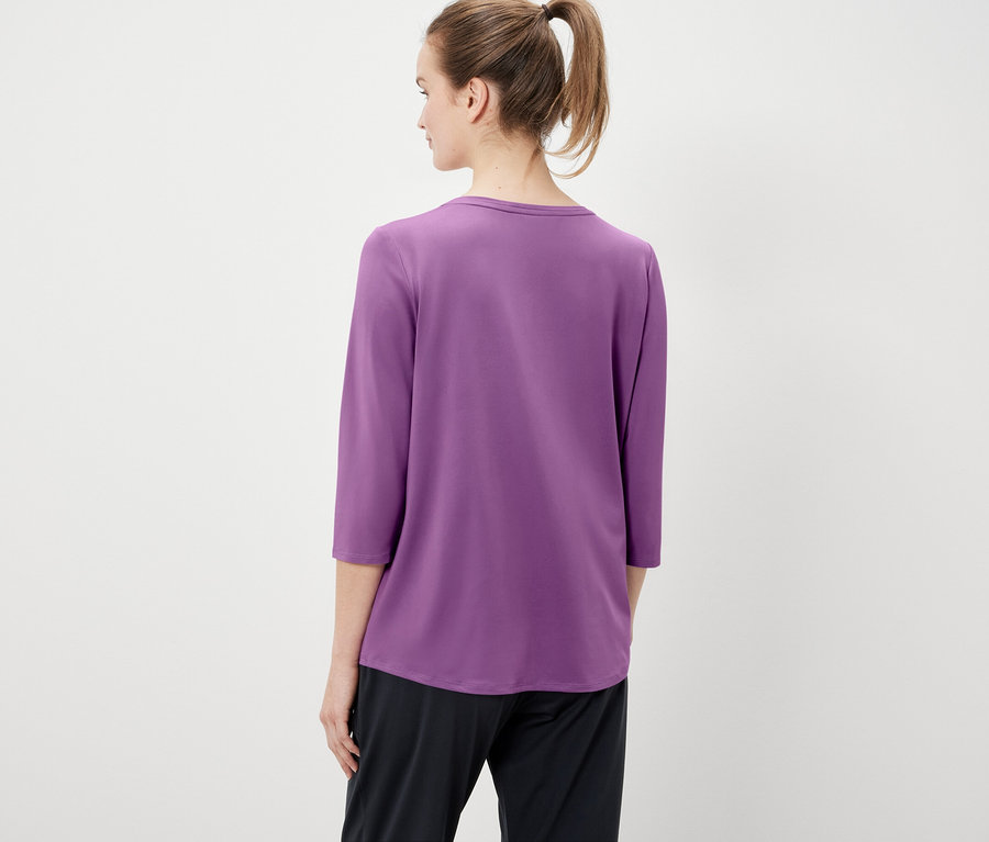 Zezadu je vidět žena s vlasy staženými do culíku, oblečená do fialového trička s tříčtvrtečními rukávy a černých kalhot.