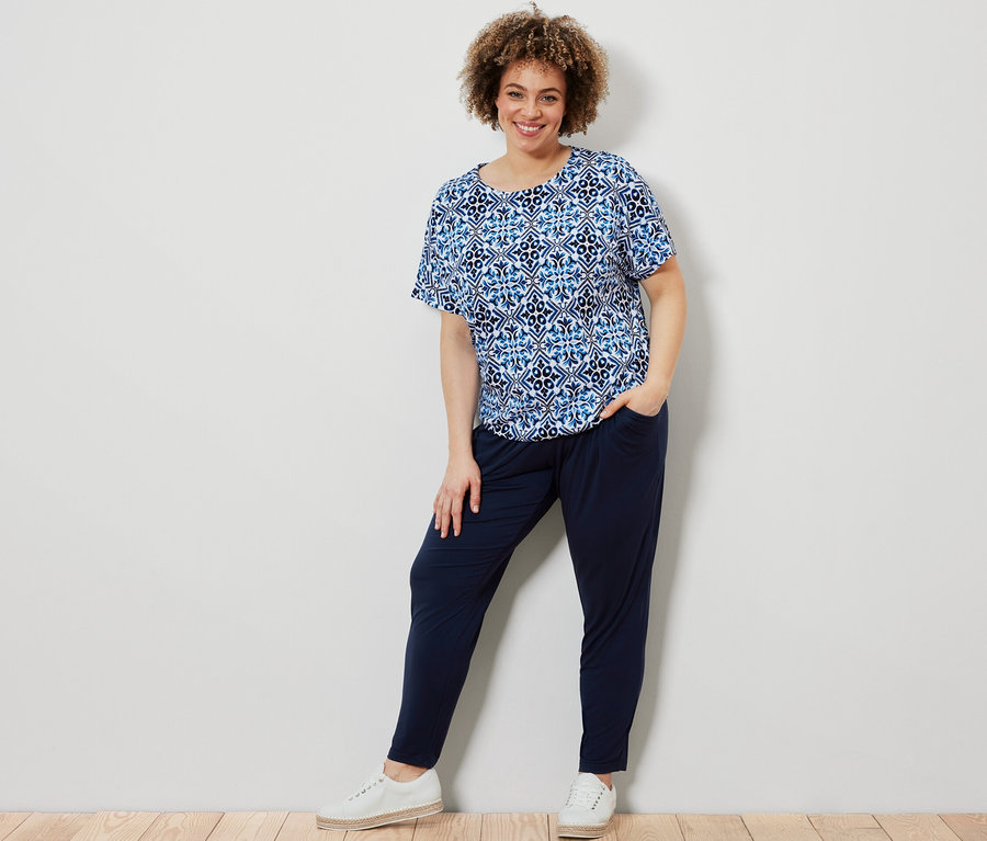 Žena pózuje v modrém halenkovém triku s potiskem a tmavě modrých žerzejových kalhotách.