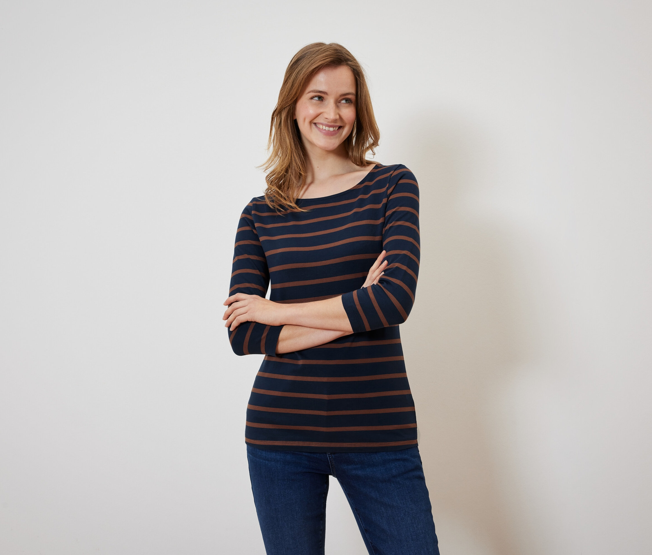Usmívající se žena pózuje s rukama zkříženýma na sobě v tričku s tmavě modrými a hnědými proužky a džínách.
