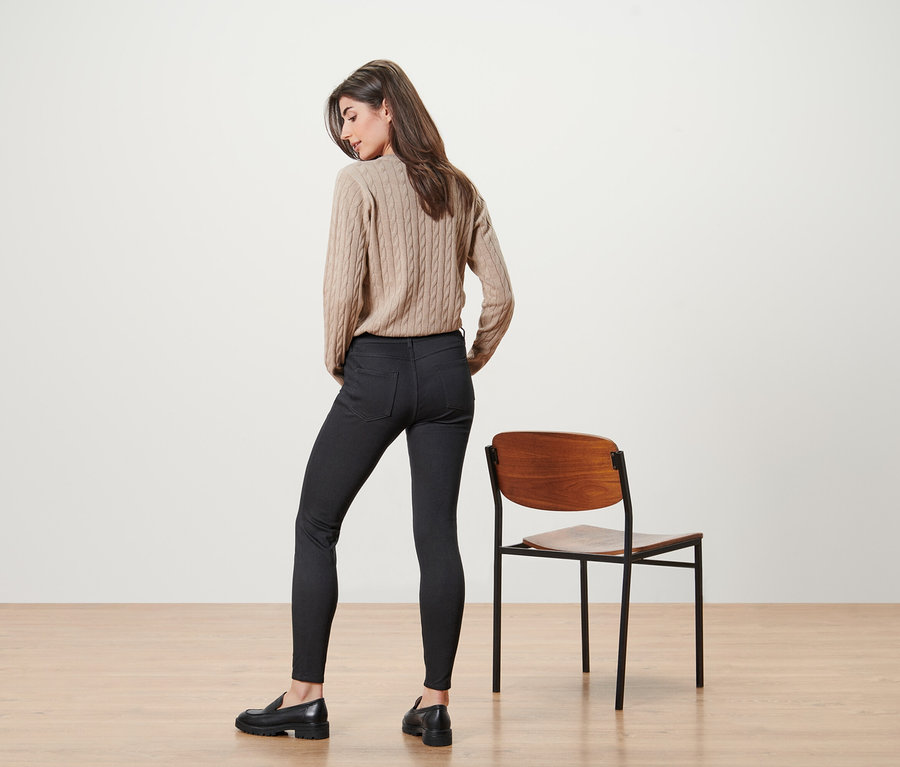 Žena pózuje v béžovém svetru a černých džínách vedle židle.