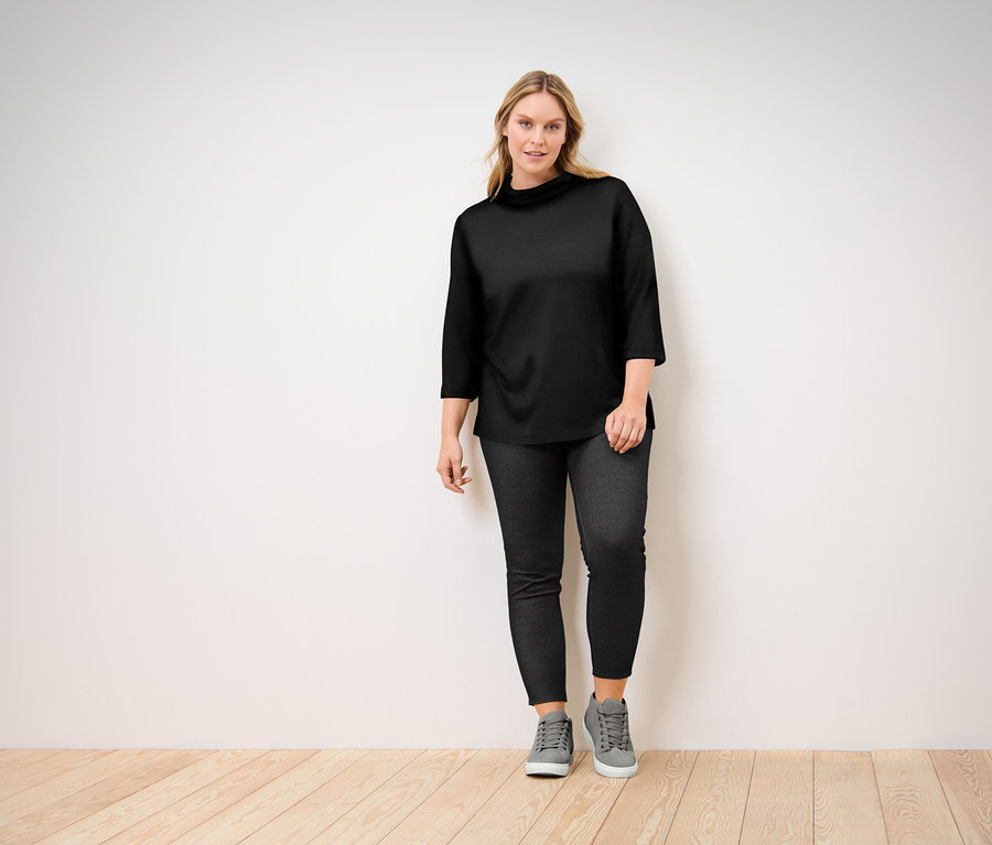 Žena pózuje v černém topu s tříčtvrtečními rukávy, černých džínách a šedých teniskách.