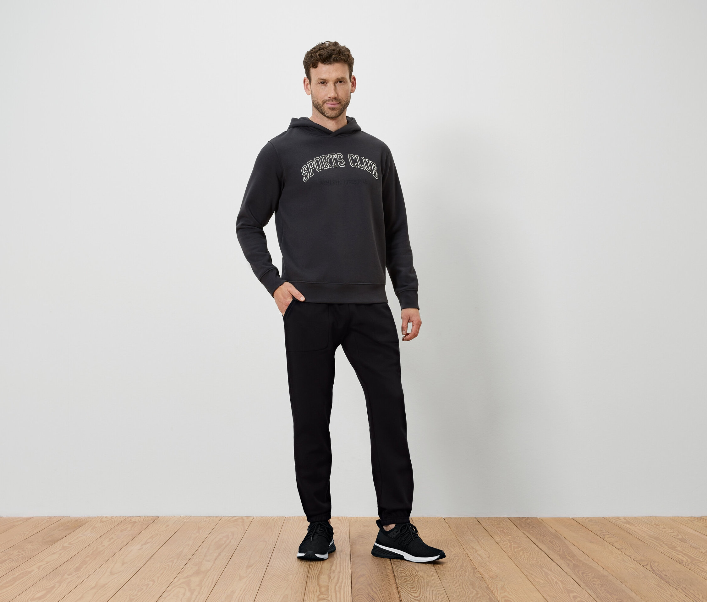 Muž stojí na dřevěné podlaze a má na sobě tmavě šedou mikinu s kapucí a černé kalhoty.