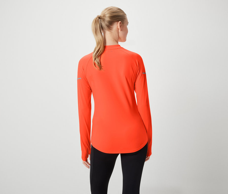 Zezadu je vidět žena s blond vlasy staženými do culíku, má na sobě oranžovou sportovní halenku s dlouhými rukávy a černé legíny.