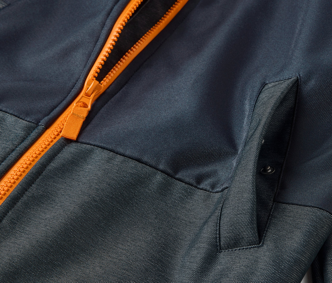 Detail dětské softshellové bundy s oranžovým zipem a kapsou.