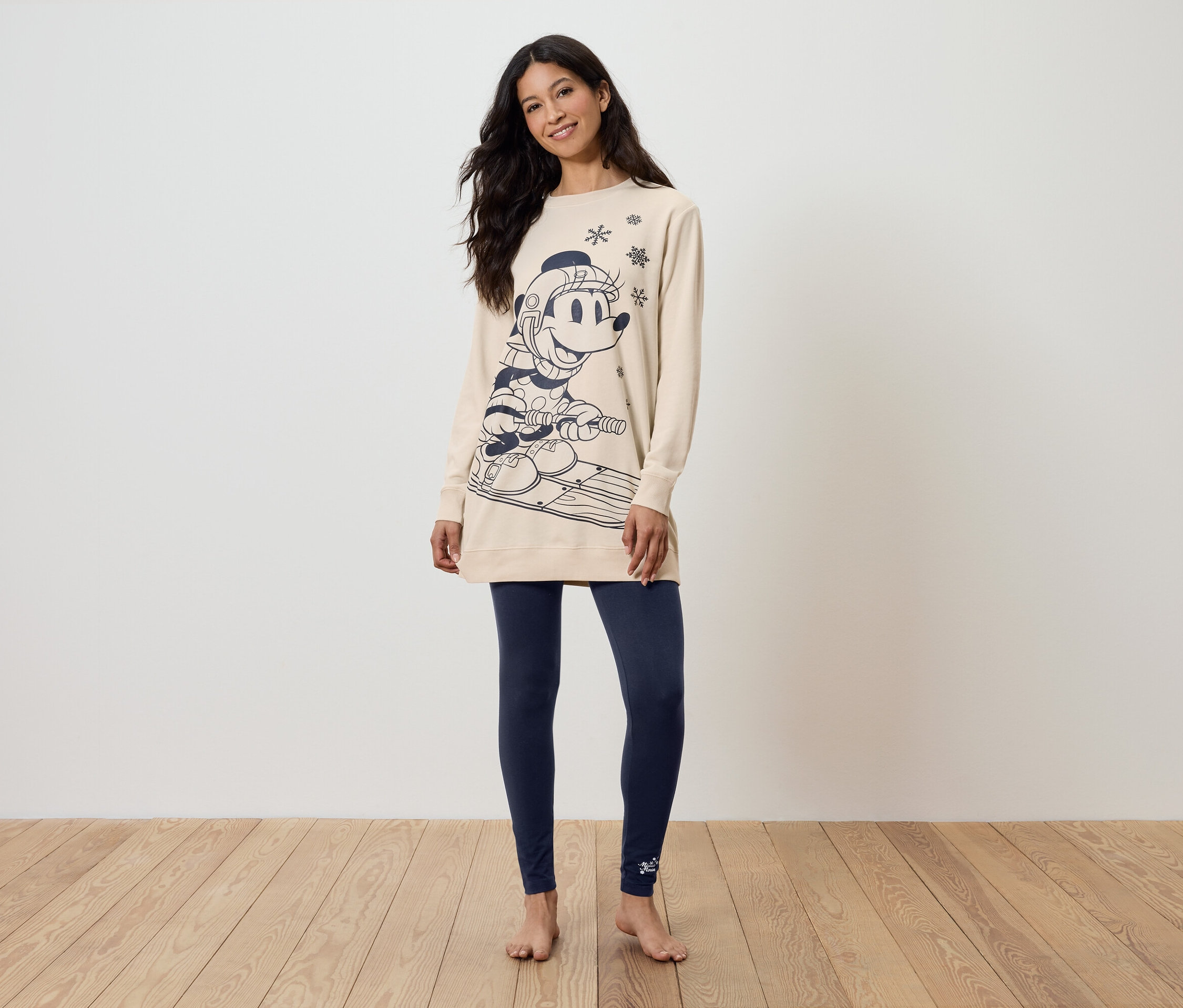 Žena stojí a má na sobě béžovou mikinu s potiskem a tmavě modré legíny.