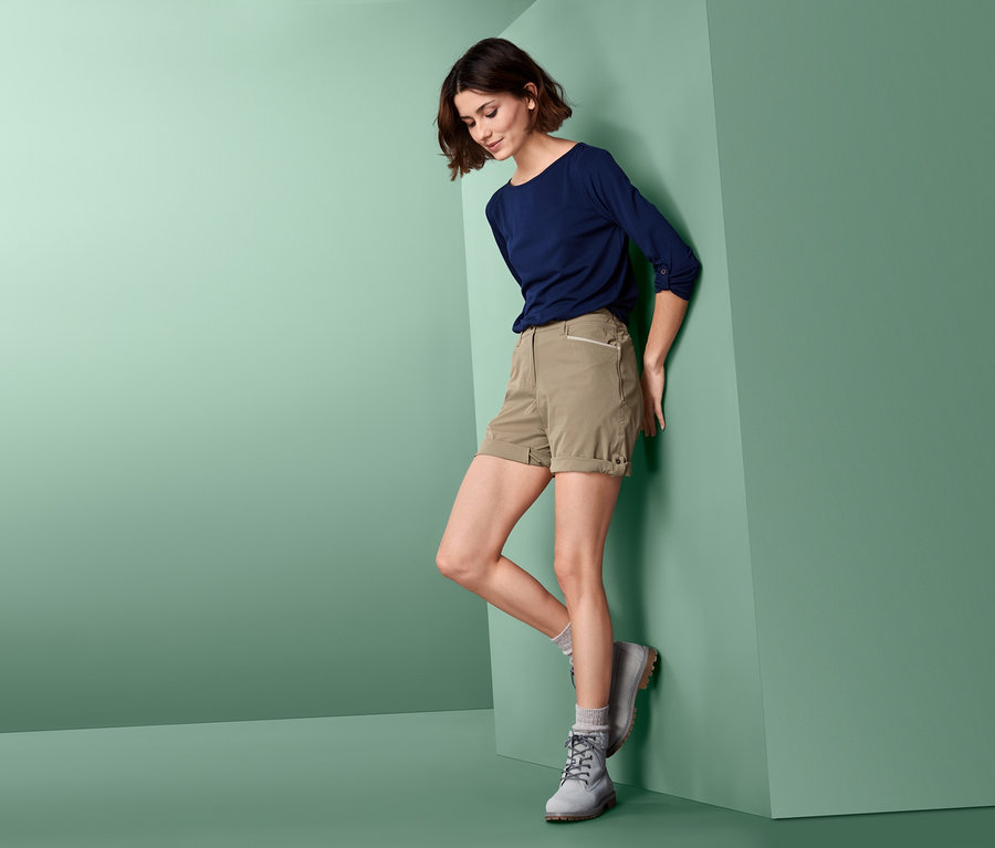 Žena opírající se o zeď nosí softshellové funkční šortky, modré tričko a šedé boty.